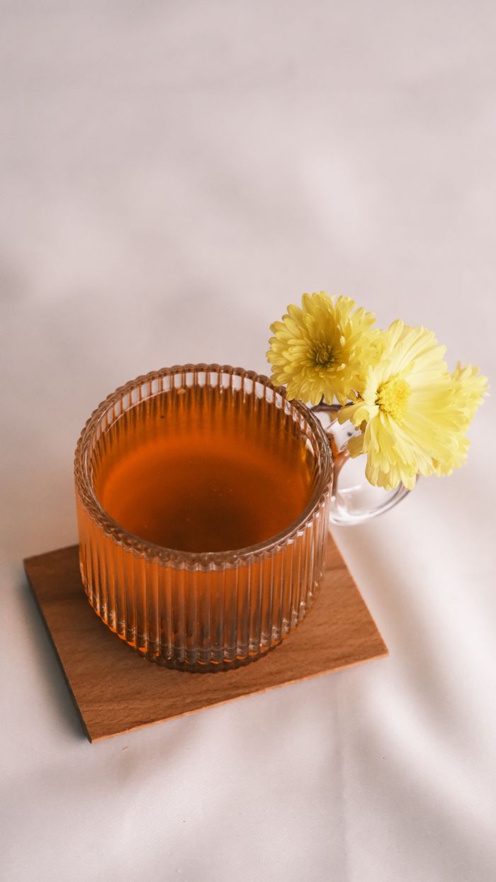 Ribbed glass mug of herbal tea with yellow flowers on wooden coaster, stylish and serene.